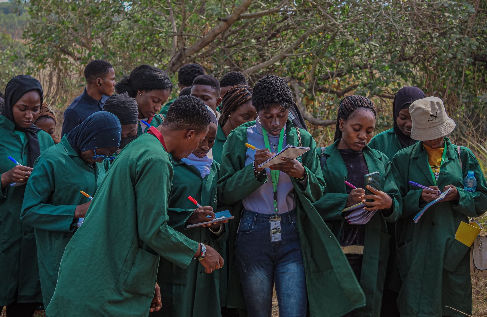African students in green coats writing notes in an outdoor setting in Nigeria.